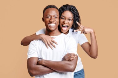 Duygusal siyahi kadın, gülümseyen erkek arkadaşına arkadan sarılıyor ve sarı stüdyo arka planında barış jesti gösteriyor. Afrika kökenli Amerikalı aşıklar neşeyle kameraya, panoramaya poz veriyorlar.