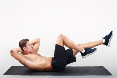 Çıplak gövdeli güçlü bir genç, yoga minderi üzerinde egzersiz yapıyor. Stüdyonun arka planında, yandan bakıyor. Çekirdek kaslarını çalıştıran yakışıklı atletik adamın tam boy portresi.