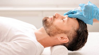 Modern kuaförde sakallı bir adamın güzellik iğnesi yaptırışının yan görüntüsü. Kozmetik uzmanı, yakışıklı bir adam için yüz doldurma işlemi için mavi lastik eldivenleri teslim etti. Yüz bakımı, erkek kozmetoloji konsepti.