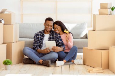 İnternetten alışveriş konsepti. Tablet bilgisayarı ve kredi kartıyla internet üzerinden ev eşyaları alan mutlu siyah çift. Genç Afrikalı Amerikalı aile internetten ev dekorasyonu satın alıyor.