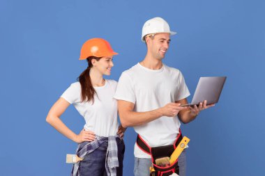 Laptop kullanan, dairelerini tamir etmek için malzeme sipariş eden şapkalı mutlu genç bir çift. Erkek ve kadın inşaat aletleri için online alışveriş yapmayı seviyorlar, mavi arka planda duruyorlar, kopyalama alanı