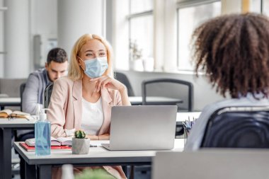 Ofisteki iş arkadaşlarının iletişimi ya da müşteri danışmanlığı ve sosyal mesafe. Koruyucu maskeli bin yıllık kadın iş yerinde bilgisayarlı, antiseptik ve bitki yetiştiren Afro-Amerikan bayanla konuşuyor.