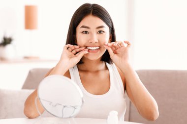 Mükemmel gülümseme, diş sigortası, temiz ağız ve ağız bakımı kavramı. Evde diş ipi kullanan, sabah rutinini yapan, kameraya bakan kendine güvenen Asyalı kadın modelin portresi.