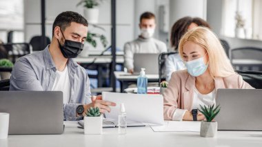 Modern ofisteki belgelerle sosyal mesafe ve çalışma. Bin yıllık koruyucu maskeli kadın ve erkek iş yerinde laptoplar ve antiseptiklerle karantina sonrası konuşuyor.