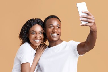 Cheerful african american couple taking selfie on mobile phone over yellow studio background, copy space. Joyful young black man and woman lovers taking photos while bonding, using smartphone