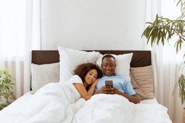 Gülümseyen çekici milenyum çifti akıllı telefon kullanıyor, birlikte yatakta uzanıyor, mutlu çift sosyal ağları kontrol ediyor, sabahları haber okuyor, genç Afrikalı Amerikalı erkek kadınlara yeni mobil uygulama gösteriyor