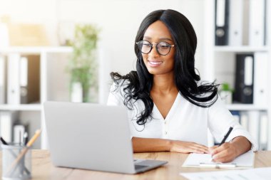 Diz üstü bilgisayarda çalışan neşeli siyahi iş kadını. İnternetteki iş toplantısında not alıyor ya da ofiste Webinar giyiyor. Mutlu Afrikalı Amerikalı sekreter bilgisayar ekranından önemli bilgiler yazıyor