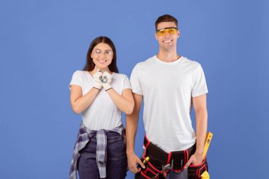 Onarıma hazır güvenlik gözlüğü takan mutlu bir çiftin portresi, Milenyum Erkek ve Kadın, Mavi Stüdyo Arkaplanı Üzerinde Yenileme Aletleri Pozisyonları, Kamerada Gülümseme, Uzay Kopyalama