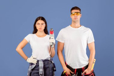 Yenilemeye hazırım. Koruyucu gözlük takan, inşaat kurutma makinesini tutan, mavi stüdyonun arka planında duran, ciddi tamirci ve tamirci bir çift portresi.