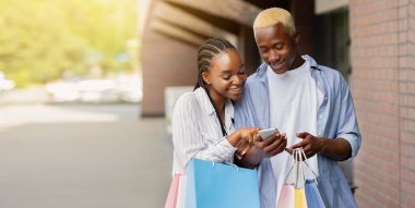 Fiyatlar ve satış izleme. Renkli çantalı Afrikalı Amerikan çifti butiğin yakınındaki akıllı telefona bakıyor, uzay boşluğunu kopyalıyorlar.
