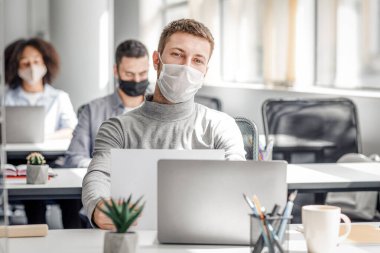 Tecritten sonra iş ve koronavirüs salgını. Koruyucu maske takan milenyum erkek çalışanı, iş yerinde koruyucu camlar ve bilgi arayışıyla evrak ve dizüstü bilgisayarla çalışıyor.