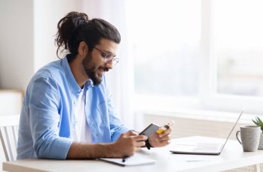 Genç Arap adam evde akıllı telefon ve kredi kartı kullanıyor, cep telefonu ve cep telefonu uygulaması ile online alışveriş yapıyor, gülümseyen doğulu adam kolay ödemelerden ve internetten satın almaktan hoşlanıyor,