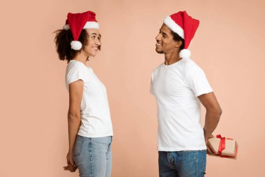 Yılbaşı ve Noel için sürpriz hediye. Gülümseyen Afrikalı genç Amerikalı kadın Noel Baba şapkalı kocadan hediye bekliyor. Arkasında kutu var, arka planda izole edilmiş, boş alan, stüdyo çekimi.