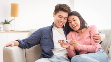 Ebeveyn olacağız. Neşeli evli Çinli çift pozitif hamilelik testi yapıyorlar. Evdeki koltukta oturup kutluyorlar ve sarılıyorlar. Aile Anları, Doğum, Mutlu Ebeveynler Bebek Bekliyor