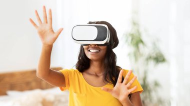 Gelecekçi ev hobileri. Sanal gerçeklik kulaklıklı Afrikalı Amerikalı kadın içeride interaktif bilgisayar oyunu oynuyor. VR gözlüklü siyahi kadın siber uzayı keşfediyor.