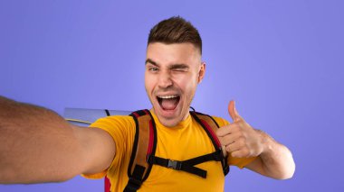 Havalı genç hippi turist ekipmanlarıyla selfie çekiyor, göz kırpıyor ve mavi stüdyo geçmişine el hareketi yapıyor. Kamp yapmadan önce komik bir fotoğrafını çeken pozitif bir yürüyüşçü.