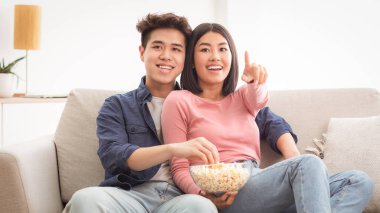 TV 'de komedi filmi seyreden, patlamış mısır yiyen hafta sonunu birlikte evde koltukta oturarak geçiren neşeli Asyalı çift. Koreli Eşler Oturma Odasında Sinemadan Hoşlanıyor
