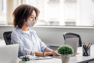 Karantinadan sonra modern ofiste müdürlük işi. Koruyucu maskeli Afrikalı Amerikalı bir kadın iş yerindeki bilgisayarında covid-19 salgını sırasında antiseptikle yazı yazıyor.