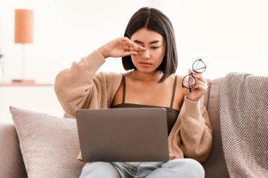 İş yorgunluğu. Uykulu Asyalı kadın göz yorgunluğu çekiyor, gözlük tutuyor, gözleri ovuyor, dizüstü bilgisayarda çalışıyor, oturma odasında kanepede oturuyor. Yaralı gözlere dokunan kadın