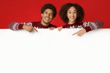 Mutlu yıllar ve kış tatili kutlamaları. Gülümseyen bin yıllık Afro-Amerikan kadın ve aynı Xmas kazağı giyen adam beyaz ilan panosu tutuyor ve kırmızı arka planda izole edilmiş parmaklarıyla ona işaret ediyor.