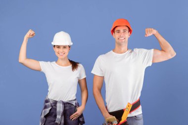 Koruyucu miğferler takmış neşeli erkek ve kadın yumruklarını kaldırıyor, kaslarını gösteriyor mavi stüdyo arka planında poz verirken kaslarını gösteriyor, güçlü işçi çift evi yenilerken eğleniyor, yer kopyalıyor.