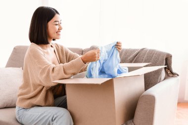 Mutlu Müşteri Konsepti. Paket kutusunu açıp açan Asyalı heyecanlı bir bayan, elinde bluz, evdeki oturma odasındaki kanepede oturuyor. Teslimat hizmetinden memnun gülümseyen genç kadın.
