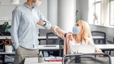 Coronavirus salgını ve hastalıklardan korunma döneminde modern bir selamlama. Koruyucu maskeli genç adam ve kadın dirsekleriyle dokunarak ofis içi boşluğa merhaba diyor.