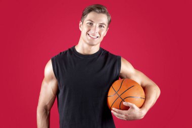 Kırmızı stüdyo arka planında top tutan mutlu basketbolcu portresi. Olumlu milenyum sokak oyuncusu gülümseyen ve kameraya bakan, turnuvaya katılmaya hazır
