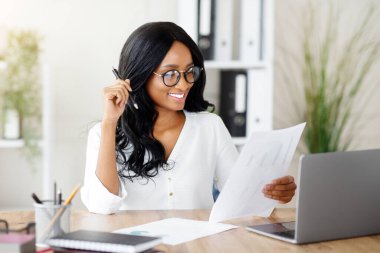 Çağdaş ofisindeki masasında doküman okuyan profesyonel bayan yöneticinin portresi. Milenyum siyahi girişimcisi iş yerinde laptopun yanında evrak işleriyle uğraşıyor.