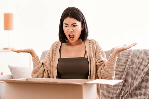 Bad Delivery Service Concept. Angry mad asian woman screaming and spreading hands after unpacking cardboard box, received damaged item, sitting on the couch, feeling upset with wrong parcel