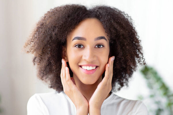 Woman touching soft cheek with clean and fresh skin with positive emotional. Cheerful young african american woman enjoying makeup and presses hands to face in living room interior at home, copy space