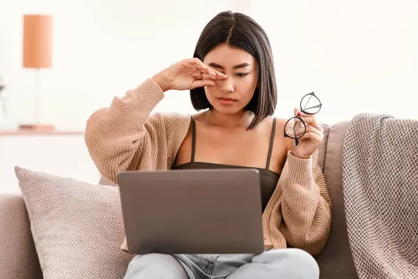 İş yorgunluğu. Uykulu Asyalı kadın göz yorgunluğu çekiyor, gözlük tutuyor, gözleri ovuyor, dizüstü bilgisayarda çalışıyor, oturma odasında kanepede oturuyor. Yaralı gözlere dokunan kadın