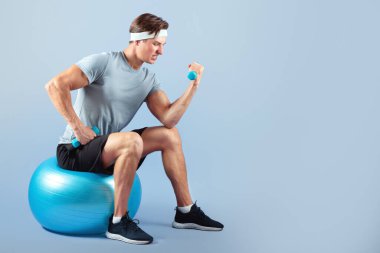 Leylak rengi stüdyo arka planında fitness topunun üzerinde otururken dambıllarla egzersiz yapan kararlı bir milenyum sporcusu. Motive olmuş, ağırlık kaldıran, aerobik yapan bir gencin uzun bir portresi.