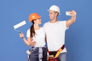 Koruyucu Şapkalı, Smartphone 'da selfie çeken, mavi arka planda poz veren, evi yenilemeye hazır neşeli tamirci çift. Alet kemeri takan adam ve boyalı paten tutan kadın.