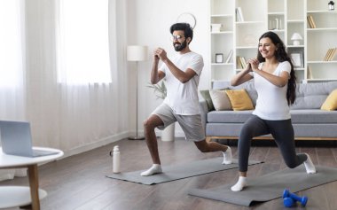 Evde antrenman. Spor yapan Hintli çift laptopun önünde kalça egzersizi yapıyor, mutlu genç doğu eşleri online dersler izliyor ve oturma odasında egzersiz yapıyor, birlikte spor yapıyorlar.