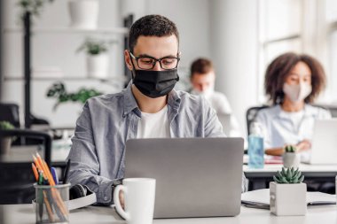 Ofis çalışanları covid-19 salgını sırasında virüs salgınından korunuyor. Gözlüklü ve koruyucu maskeli genç hippi dizüstü bilgisayarda çalışıyor. İç mekanlarda iş arkadaşları var, ücretsiz.