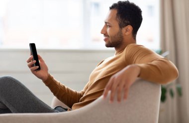 Oturma odasındaki koltukta oturan gülümseyen Arap 'ın portresi. Havalı adam internette geziniyor, internette sörf yapıyor ya da video seyrediyor, uygulama kullanıyor, boş alan