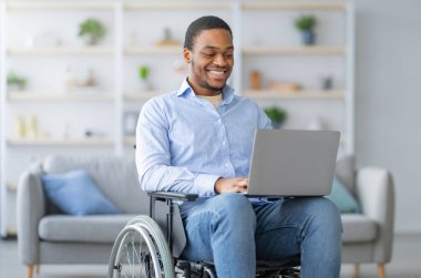 Evdeki uzaktan iş için dizüstü bilgisayar kullanan tekerlekli sandalyedeki neşeli, engelli siyahi bir adamın portresi. Fiziksel engelli mutlu zenci adam covid salgını sırasında online çalışıyor.