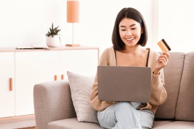 Çevrimiçi Ödeme Kavramı. Gülümseyen Asyalı kadın internet üzerinden alışveriş yapıyor, bilgisayar kullanıyor, kredi kartı tutuyor ve gösteriyor. Dizüstü bilgisayarı olan tatmin olmuş bayan koltukta oturuyor, boş yer var.