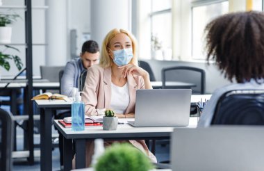 Ofisteki iş arkadaşlarının iletişimi ya da müşteri danışmanlığı ve sosyal mesafe. Koruyucu maskeli bin yıllık kadın iş yerinde bilgisayarlı, antiseptik ve bitki yetiştiren Afro-Amerikan bayanla konuşuyor.