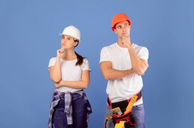 Yenileme Planlama. Koruyucu şapkalı ve günlük kıyafetleriyle stüdyoda mavi arka planda dikilen, ev tamirini düşünürken çenesine dokunan, uzay kopyası yapan düşünceli genç bir çiftin portresi.