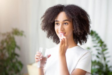 Diyet, beslenme, sağlıklı yaşam tarzı. Mutlu gülümseyen Afro-Amerikalı kadın morina yağlı hap ya da vitamin alıyor. Omega-3 ve modern yaşam ortamında sabahları elinde bir bardak taze su tutuyor.