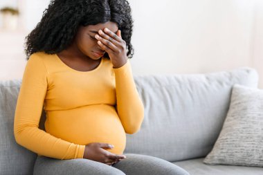 Siyah hamile kadın evde baş ağrısı çekiyor, koltukta oturuyor, alnına ve karnına dokunuyor. Afro-Amerikalı bir bayanın migreni tuttu, evde tek başına kaldı, boşluğu kopyaladı.