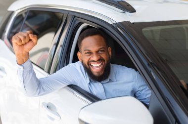 Yeni arabasında poz veren, yumruklarını kaldıran ve pencereden neşeyle bağıran heyecanlı siyahi adam bayilik merkezindeki modern araba alımını kutluyor.