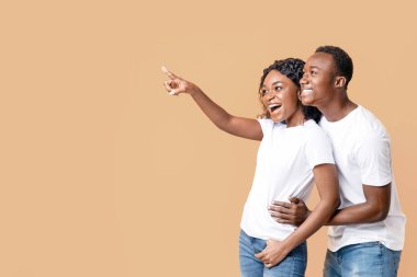 Neşeli Afro-Amerikalı çift gülümsüyor ve neşeli bir şekilde boş alanı işaret ediyor, sarı stüdyo arka planı. İlan veren mutlu siyah erkek ve kadın, kopya alanı olan panorama