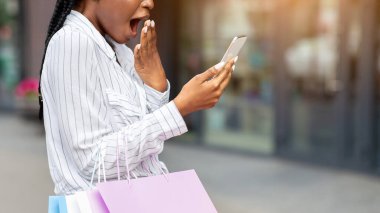 Alışveriş indirimi ya da son satış. Şok olmuş Afro-Amerikalı kadın ağzını kapatıyor ve akıllı telefona bakıyor, elinde kağıt paketler, açık hava, panorama, boş yer tutuyor.