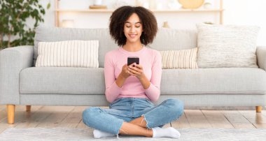 Evde online sohbet, internette sörf ve kendini izole etme üzerine yeni bir iletişim uygulaması. Gülümseyen Afro-Amerikan kadın akıllı telefonuyla mesaj yazıyor, oturma odasında yerde oturuyor.