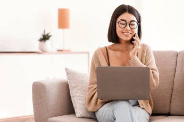 İletişim Konsepti. Gözlüklü, akıllı telefonla konuşan, dizüstü bilgisayar kullanan neşeli Asyalı kadının portresi. Modern dairesindeki oturma odasında koltukta oturan gülümseyen bayan.