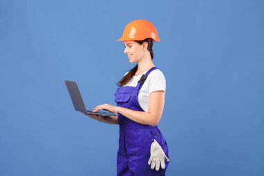 Güzel Genç Kadın Bakım İşçisi Laptop kullanıyor, İnşaat Malzemelerini İnternetten sipariş ediyor. Bayan Hardhat Onarım Aletleri Yapıyor İnternetten alışveriş yapıyor, Mavi arka planda duruyor, Boşluğu Kopyala