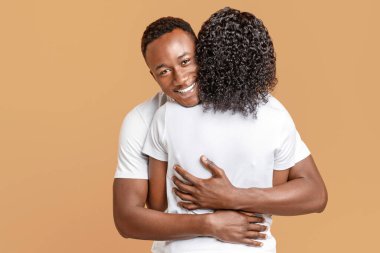 Happy black guy hugging his girlfriend, yellow studio background. African american young couple in white t-shirts cuddling, expressing feelings and emotions. Love, family, relationships concept